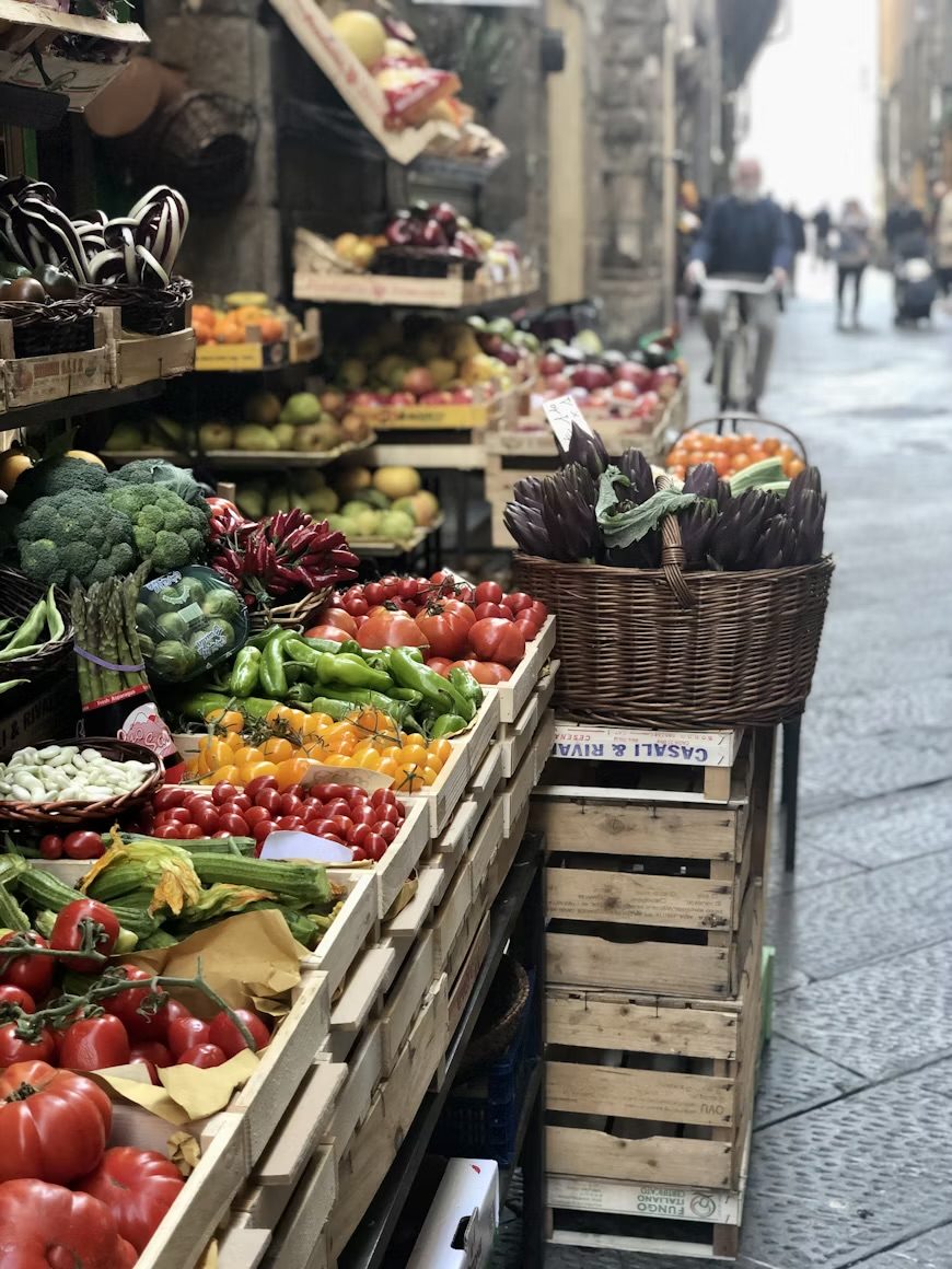 Gemüsestand in einer Gasse: Tomaten und Peperoni in Holzkisten, Artischocken im Korb