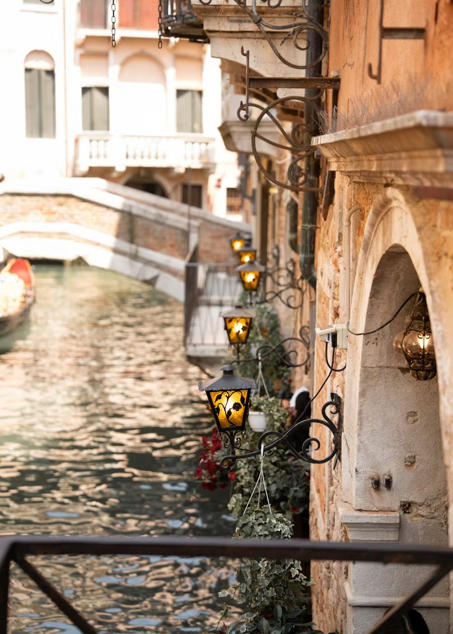 Schmaler Kanal in Venedig, leuchtende Laternen an der Hauswand, dahinter eine Brücke